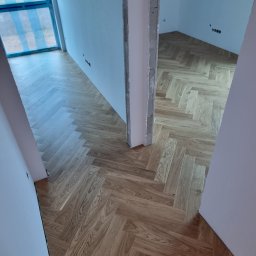 VEBARO Bartosz Nyga - Ułożona podłoga z paneli w jodełkę w jasnym odcieniu drewna, widoczna z perspektywy wejścia do pomieszczenia. Ściany w kolorze białym, surowy stan wykończenia.