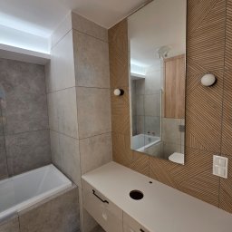 VEBARO Bartosz Nyga - Nowoczesna łazienka z beżowo-szarą glazurą, drewnopodobną ścianą, lustrem i białą szafką pod umywalkę. Widoczna wanna. Górne oświetlenie LED.