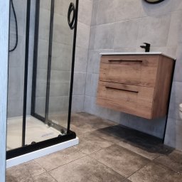 VEBARO Bartosz Nyga - Nowoczesna łazienka z kabiną prysznicową typu walk-in z czarnymi ramami, drewnianą szafką pod umywalką i okrągłym lustrem na szarej, płytkowej ścianie.