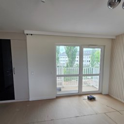 VEBARO Bartosz Nyga - Jasne wnętrze po remoncie: zabudowa szafy z czarnymi frontami, panele ścienne, okno balkonowe z widokiem na zieleń. Na podłodze płyty przygotowane do układania paneli.