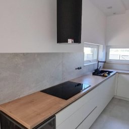 VEBARO Bartosz Nyga - Nowoczesna kuchnia z białymi szafkami, drewnianym blatem i cylindrycznym czarnym okapem. Widoczna płyta indukcyjna i szklana witryna na wino.