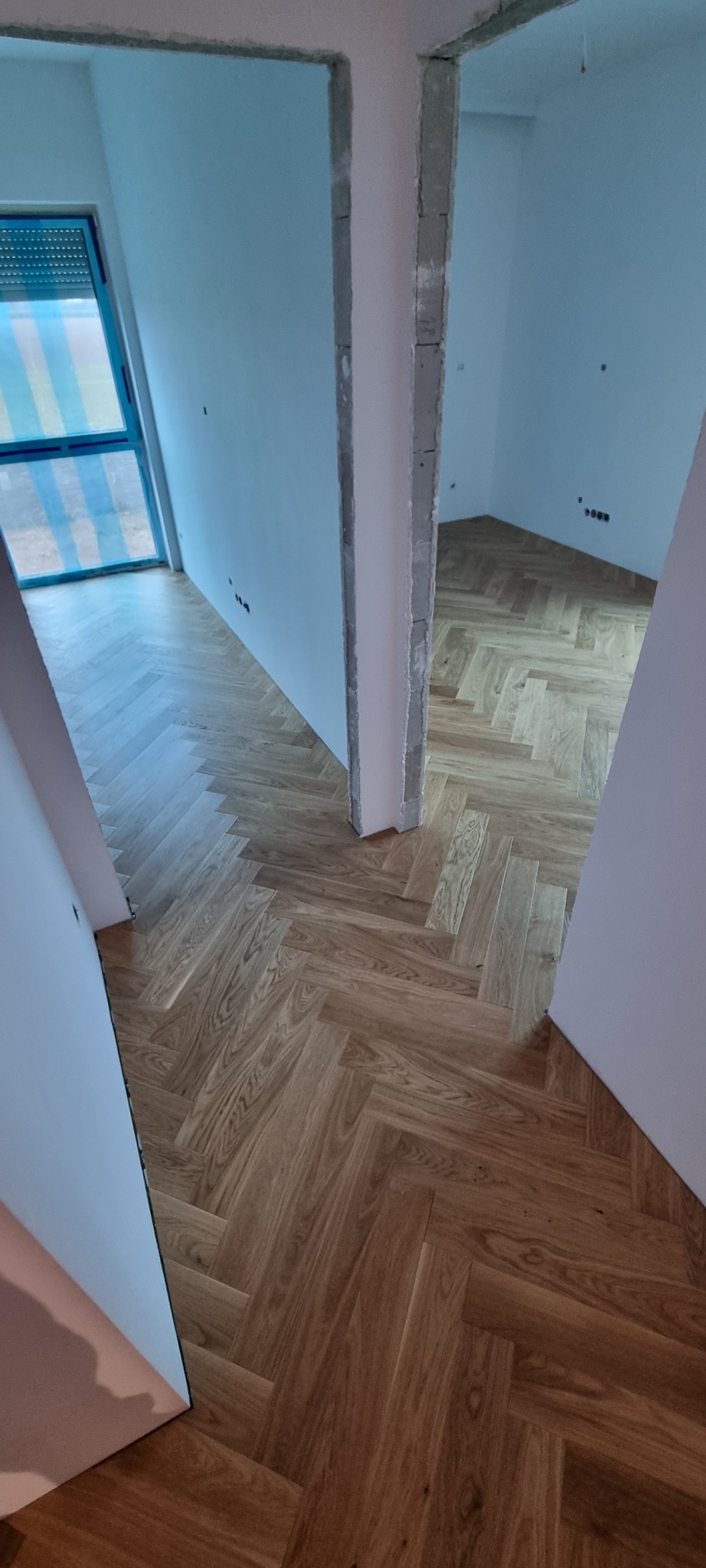 Ułożona podłoga z paneli w jodełkę w jasnym odcieniu drewna, widoczna z perspektywy wejścia do pomieszczenia. Ściany w kolorze białym, surowy stan wykończenia.