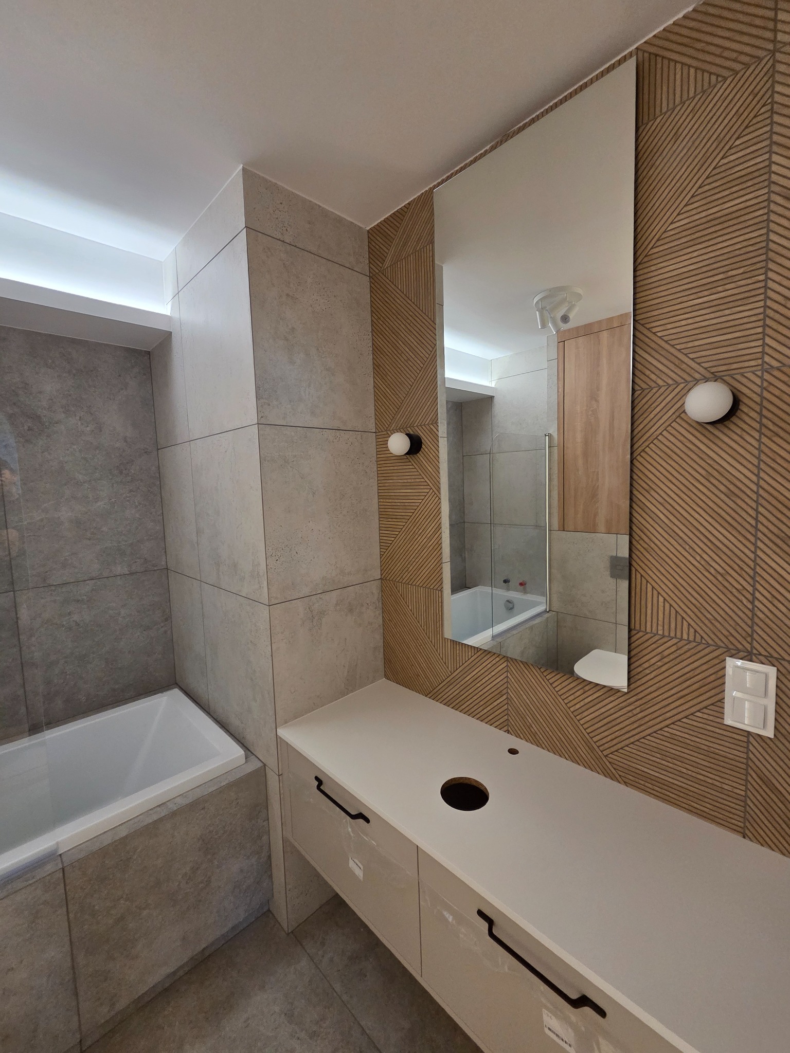 Nowoczesna łazienka z beżowo-szarą glazurą, drewnopodobną ścianą, lustrem i białą szafką pod umywalkę. Widoczna wanna. Górne oświetlenie LED.