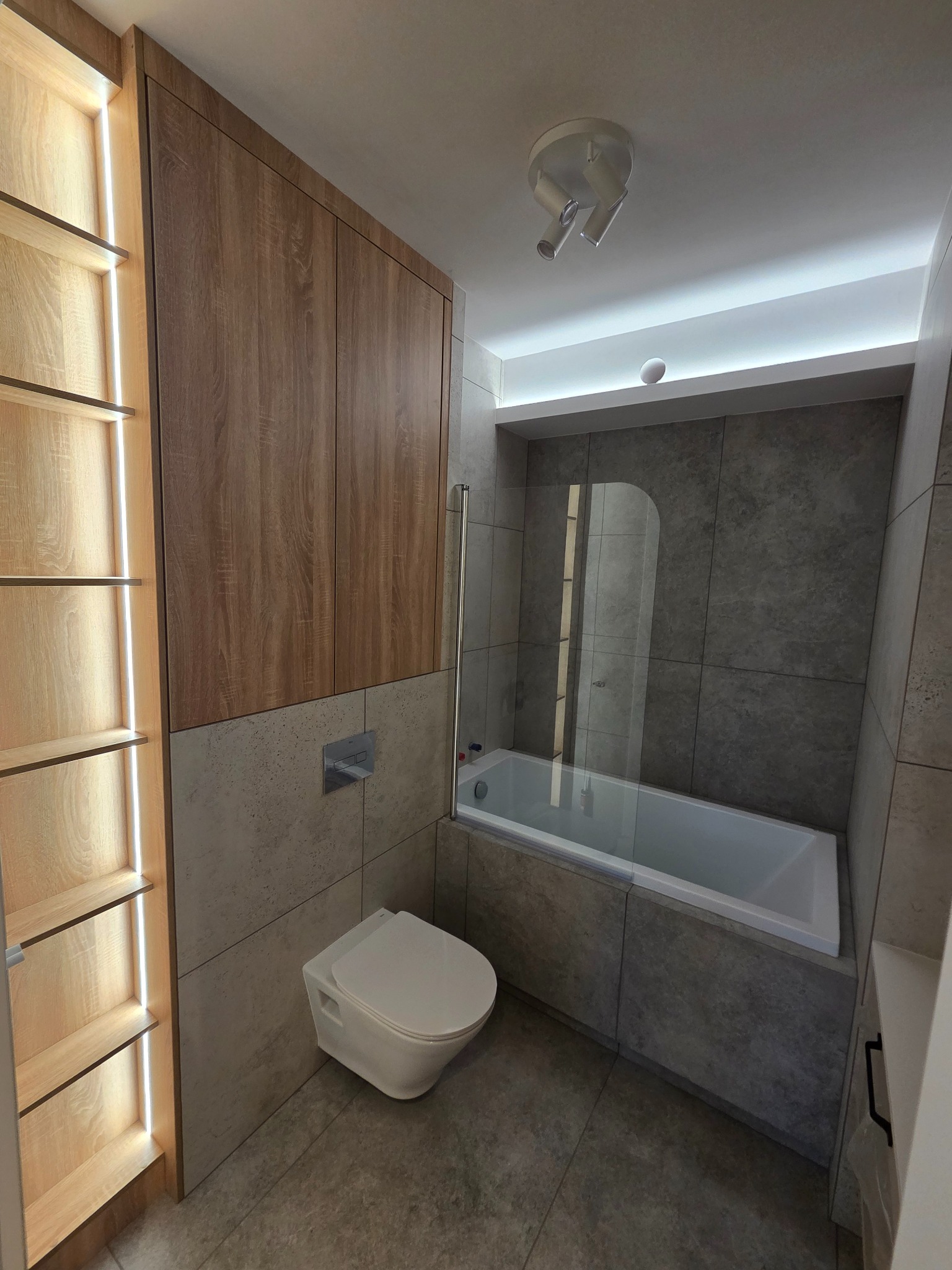 Nowoczesna łazienka z szarą glazurą, zabudową drewnianą z oświetleniem LED, białą armaturą i wanną z parawanem. Minimalistyczny design.
