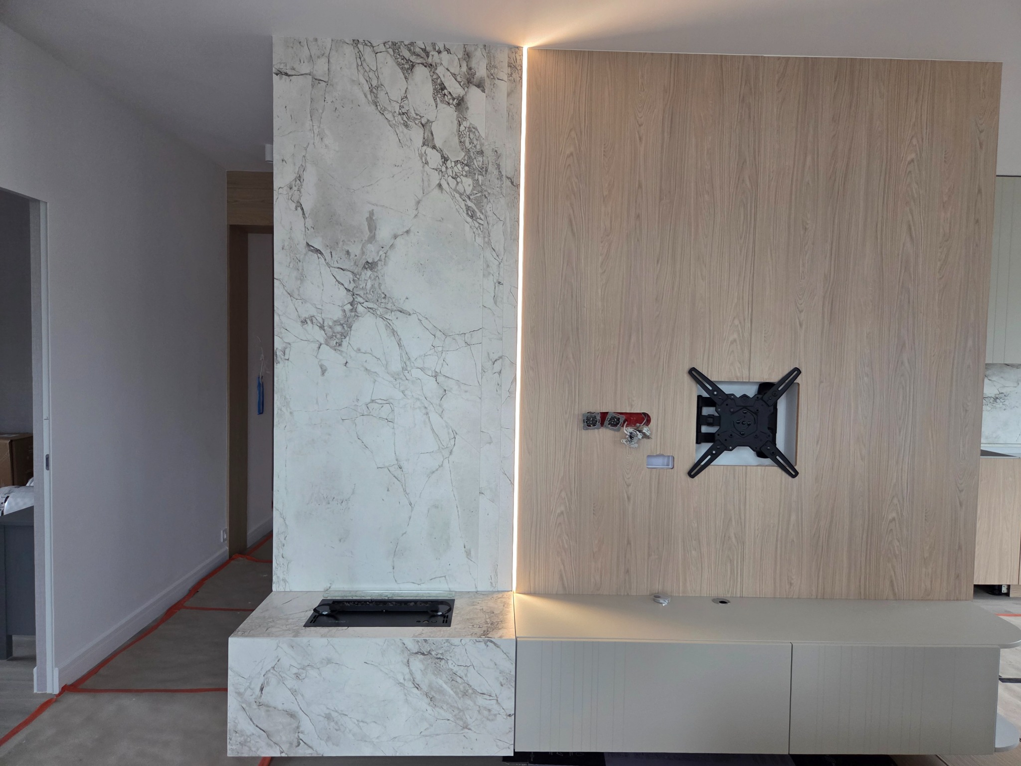 Wykończenie ściany: imitacja marmuru, drewno, podświetlenie LED. Zamontowany uchwyt TV.  Częściowo widoczne zabezpieczenia podłogi na czas prac.