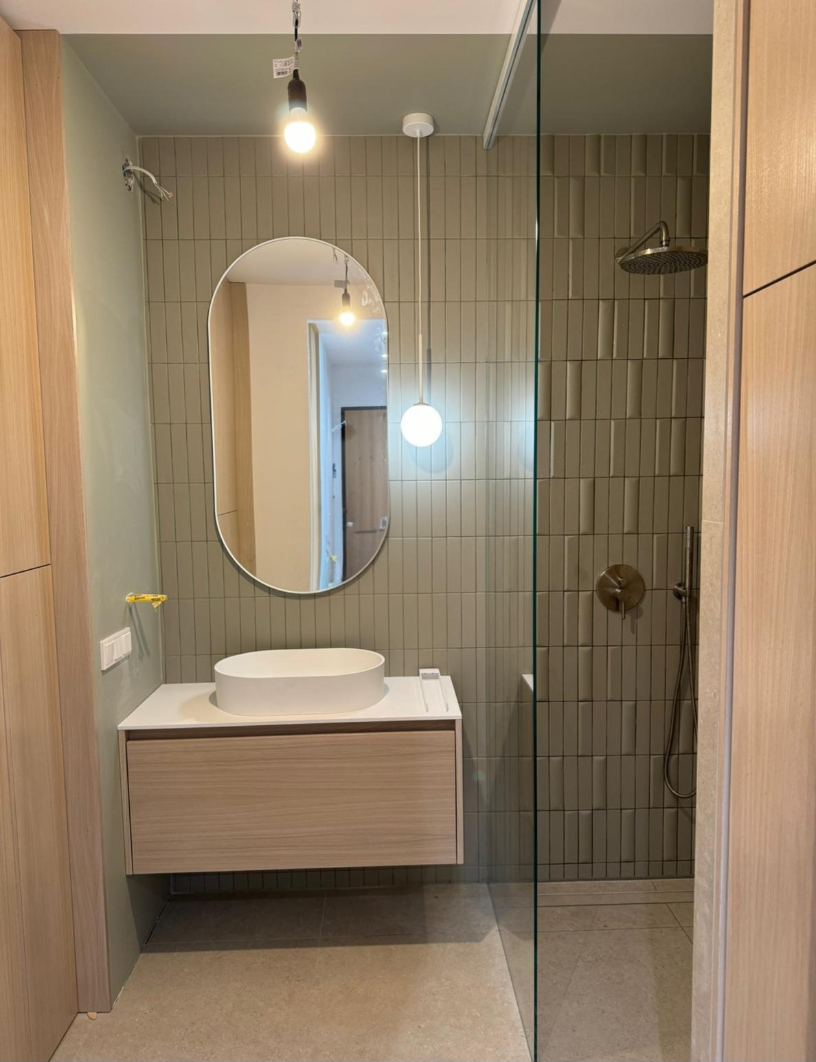Nowoczesna łazienka z owalnym lustrem, wiszącą szafką z umywalką i kabiną prysznicową wyłożoną pionowymi, beżowymi płytkami. Minimalistyczne oświetlenie.