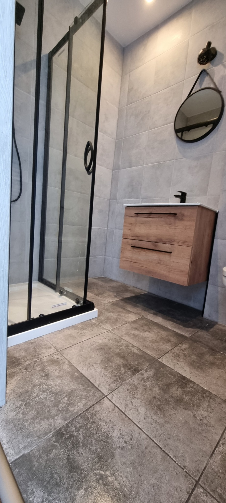 Nowoczesna łazienka z kabiną prysznicową typu walk-in z czarnymi ramami, drewnianą szafką pod umywalką i okrągłym lustrem na szarej, płytkowej ścianie.