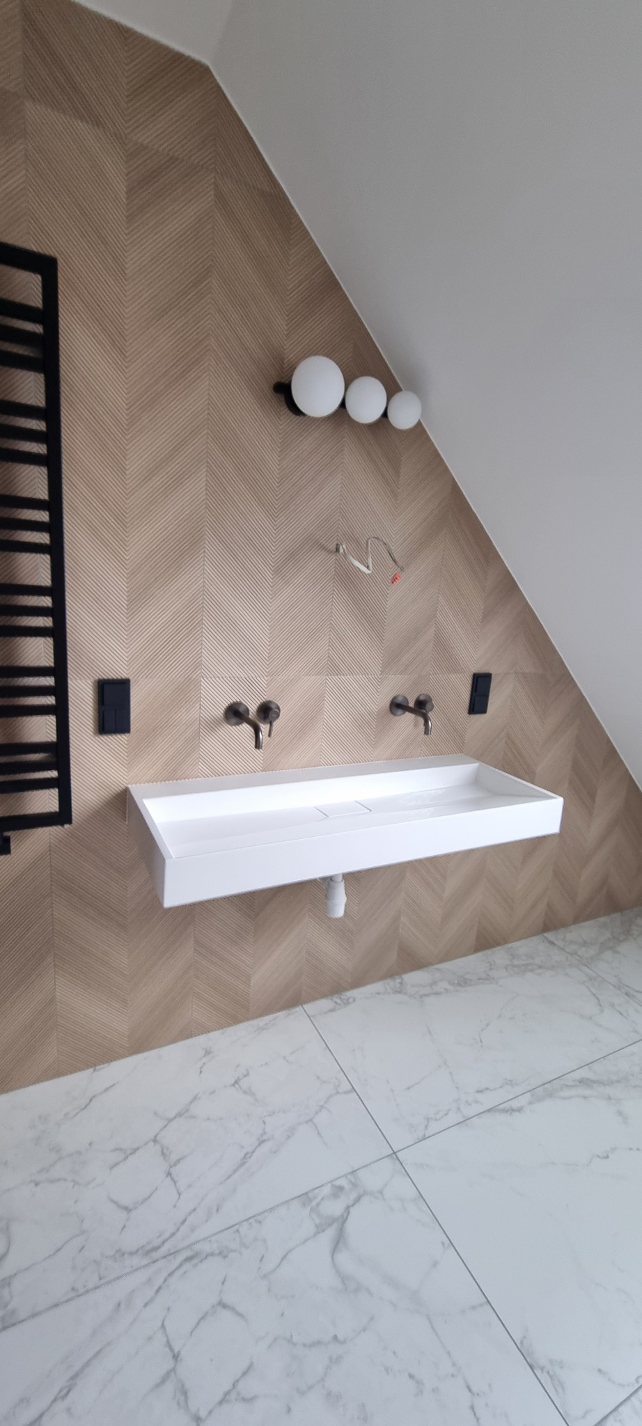 Nowoczesna łazienka na poddaszu z białą umywalką nablatową, bateriami w kolorze brązu i grzejnikiem drabinkowym na ścianie wyłożonej płytkami w jodełkę.