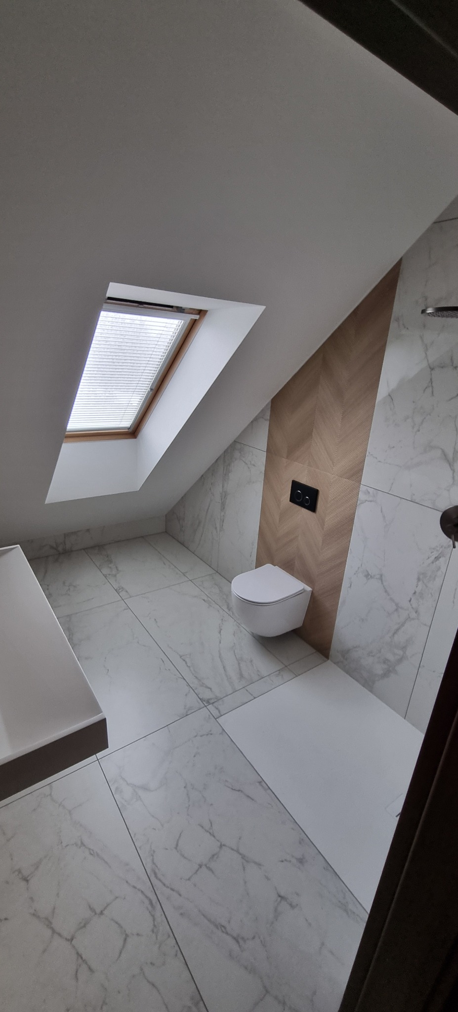 Nowoczesna łazienka na poddaszu z oknem dachowym, marmurowymi płytkami i drewnianym akcentem na ścianie. Wisząca toaleta i minimalistyczna umywalka.