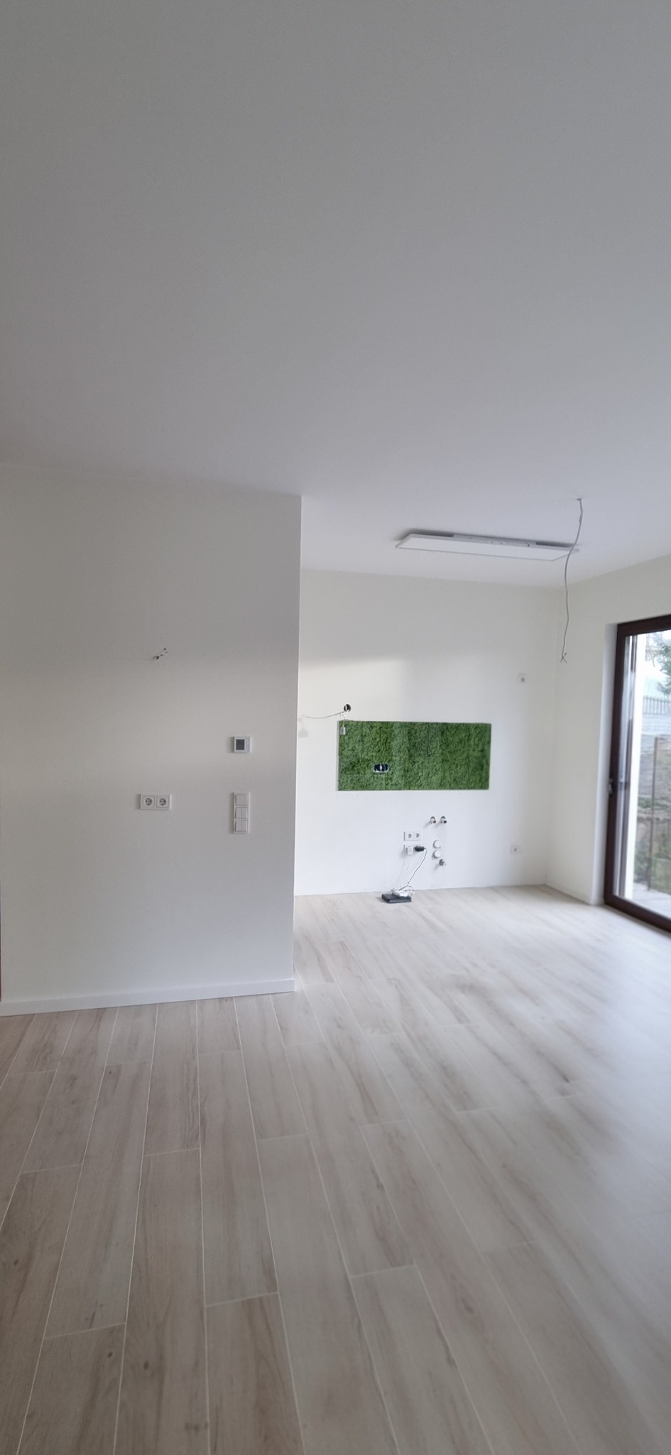 Jasne panele podłogowe imitujące drewno w przestronnym, odnowionym pokoju z białymi ścianami i zielonym panelem dekoracyjnym z mchu.