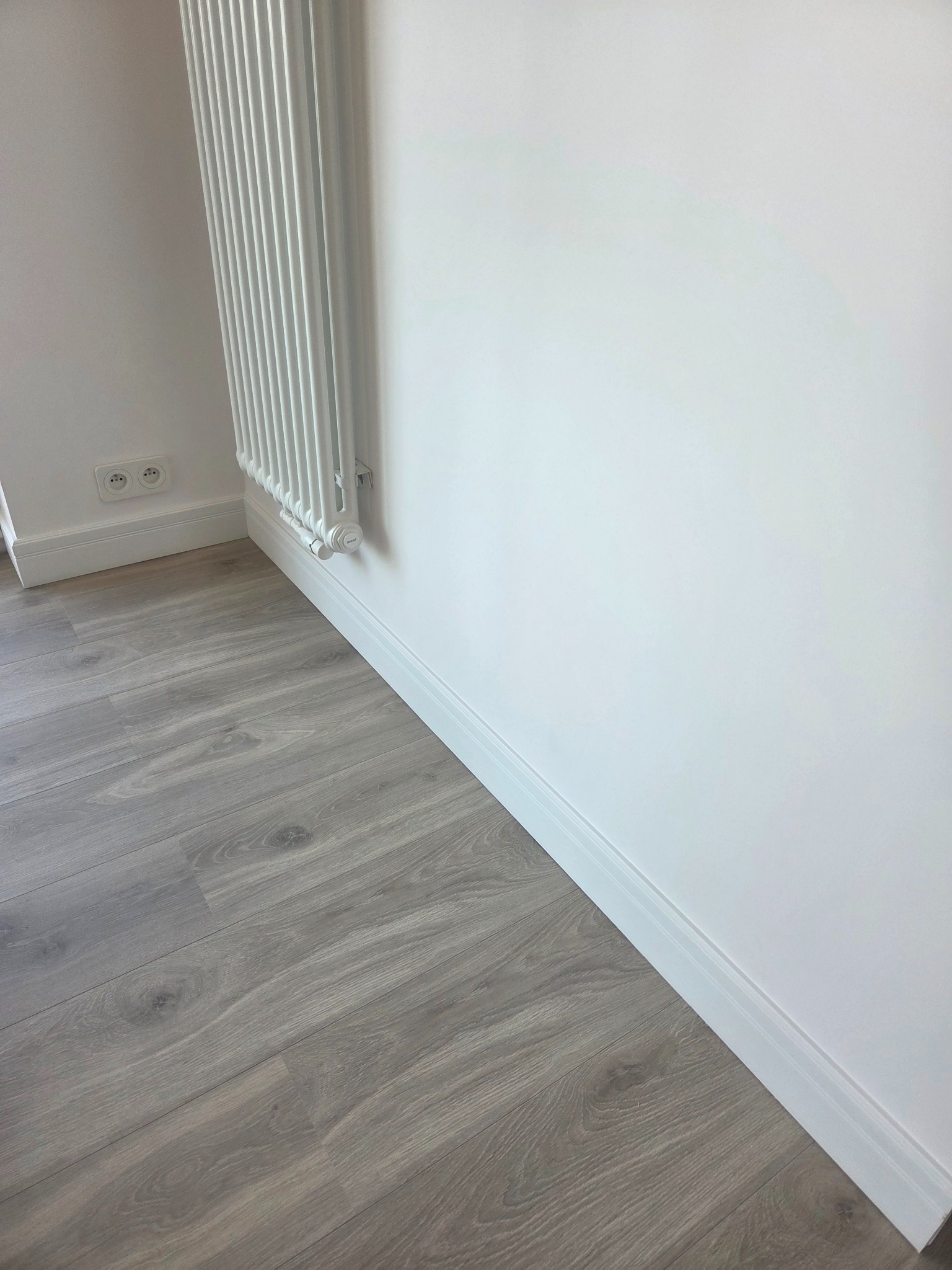 Jasne panele podłogowe i białe ściany z grzejnikiem w Dąbrowie. Wykończenie wnętrza z listwami przypodłogowymi i gładką powierzchnią ściany.