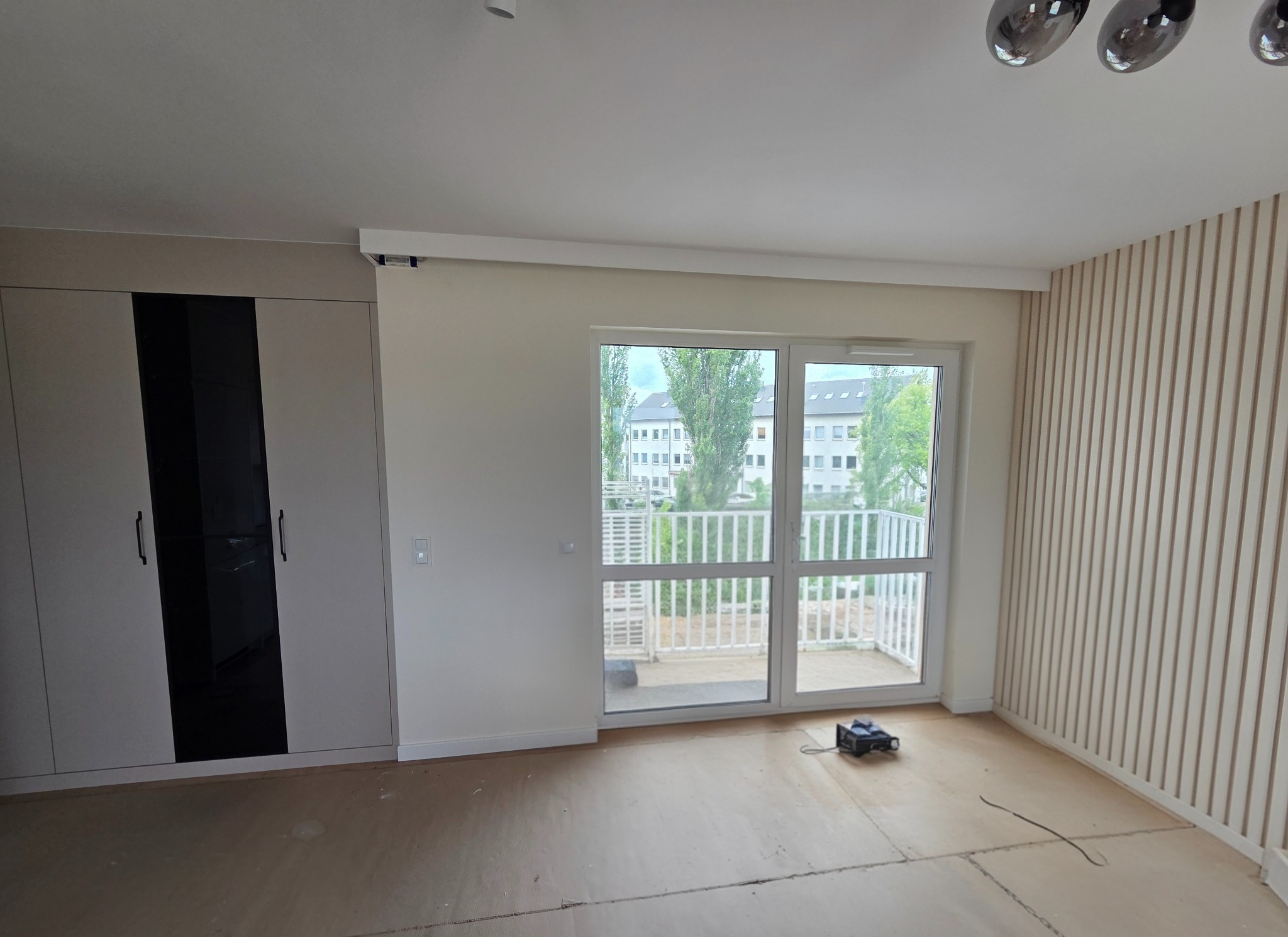 Jasne wnętrze po remoncie: zabudowa szafy z czarnymi frontami, panele ścienne, okno balkonowe z widokiem na zieleń. Na podłodze płyty przygotowane do układania paneli.