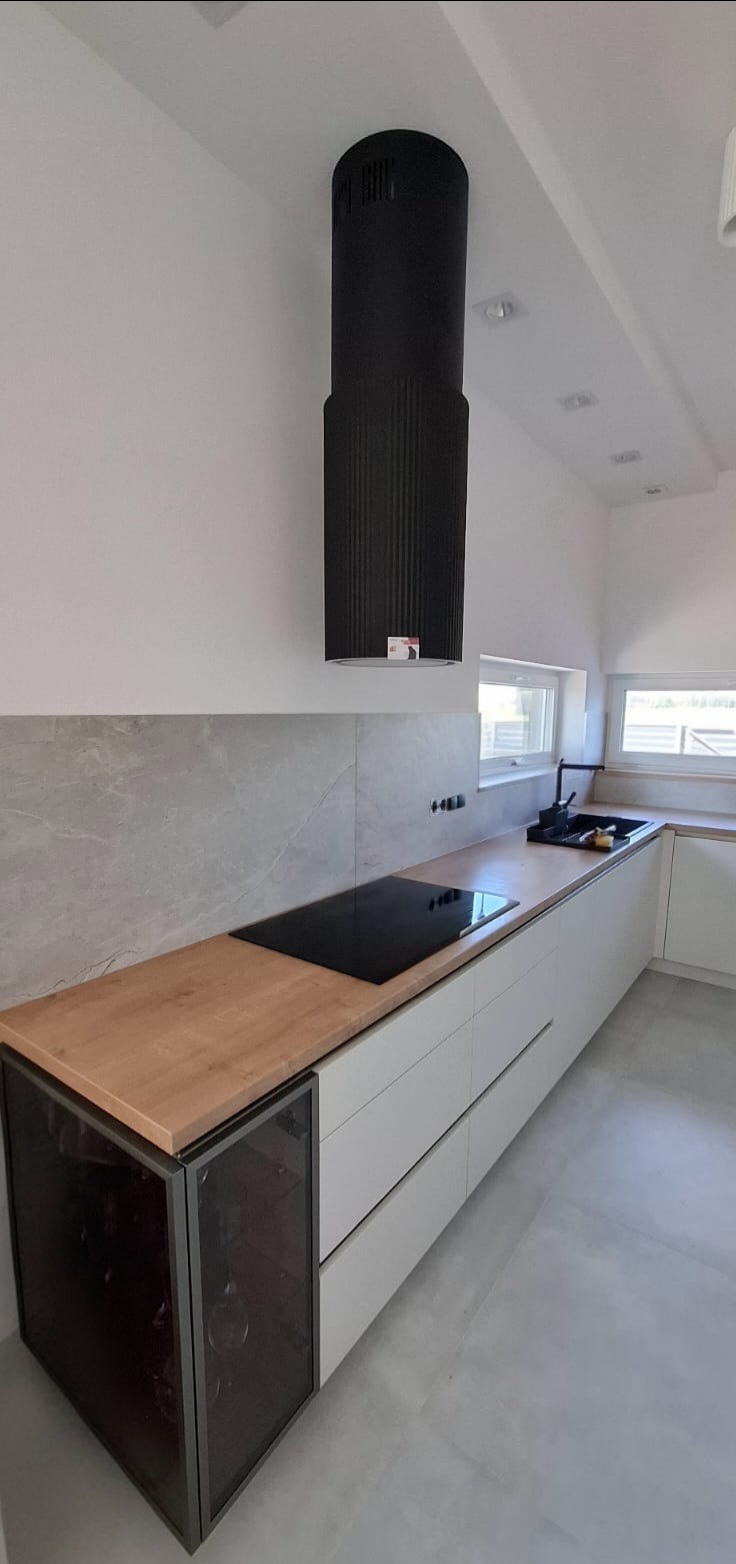 Nowoczesna kuchnia z białymi szafkami, drewnianym blatem i cylindrycznym czarnym okapem. Widoczna płyta indukcyjna i szklana witryna na wino.