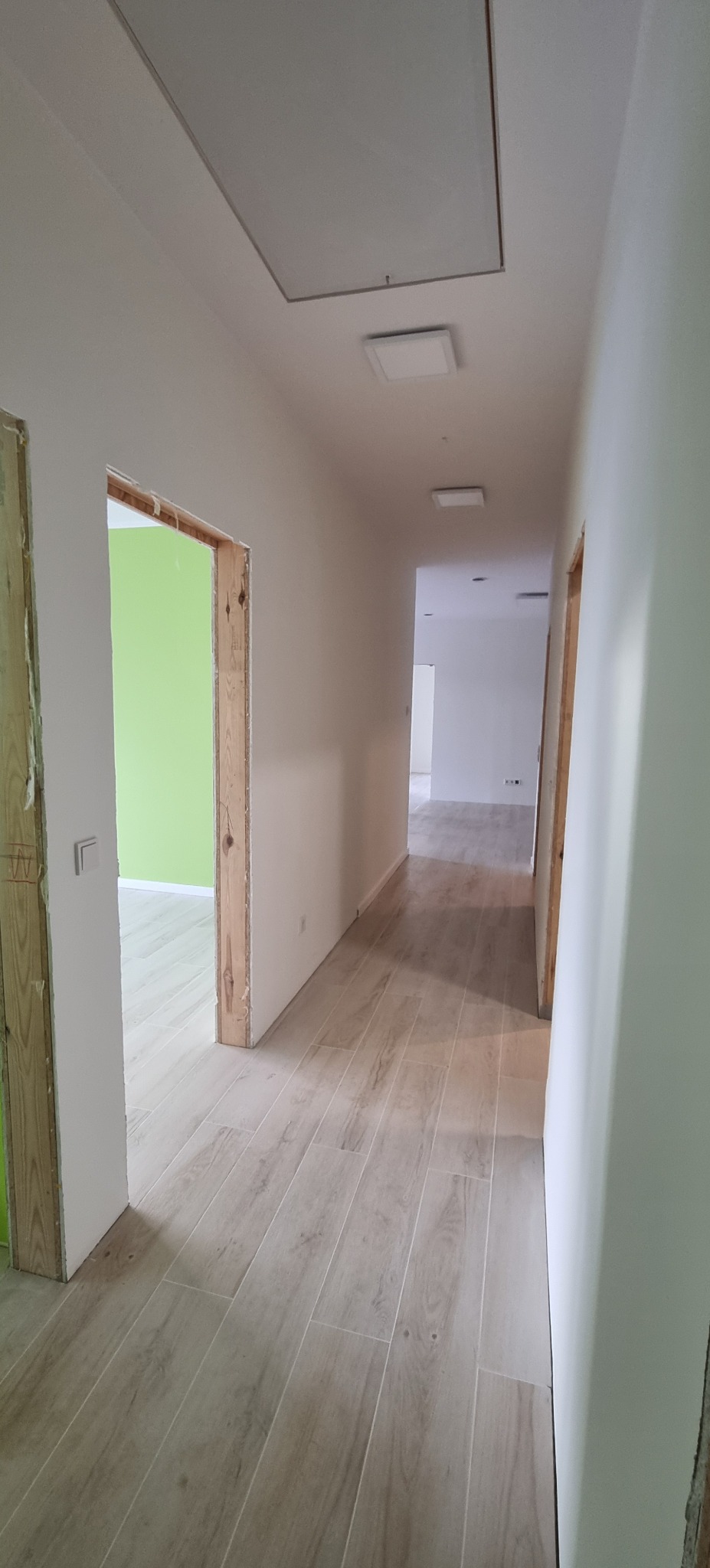 Korytarz po remoncie z drewnopodobną podłogą, świeżo pomalowanymi ścianami na biało i surowymi ościeżnicami drzwiowymi. Widok na zieloną ścianę w głębi.