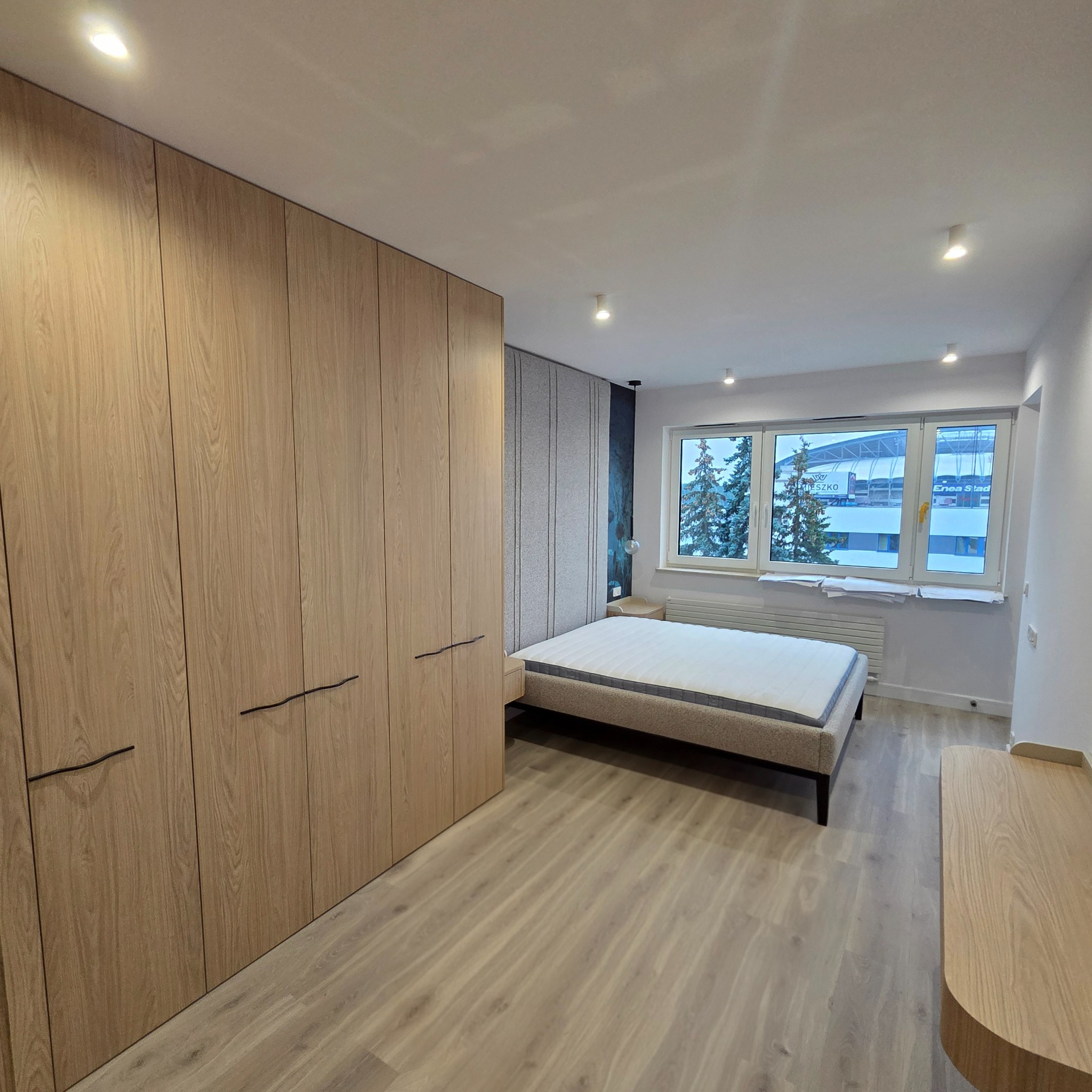 Nowoczesna sypialnia po remoncie: drewniana podłoga, zabudowa szafy z jasnego drewna, łóżko i widok na stadion przez okno. Minimalistyczny design.