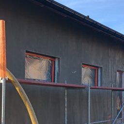 MARIUSZ SADŁOCHA - WIKBUD - Szara elewacja domu w trakcie prac wykończeniowych. Okna zabezpieczone folią, widoczne rusztowania i pomarańczowy wąż do maszyny tynkarskiej.