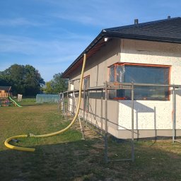 MARIUSZ SADŁOCHA - WIKBUD - Ocieplanie domu styropianem, widoczne rusztowania i żółty wąż do podawania kleju. Okna zabezpieczone folią. Prace w toku, widok z zewnątrz, słoneczny dzień.