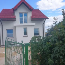 Biały dom z czerwoną dachówką, widziany zza zielonego ogrodzenia i bujnej roślinności. Błękitne niebo z chmurami odbija się w oknach.