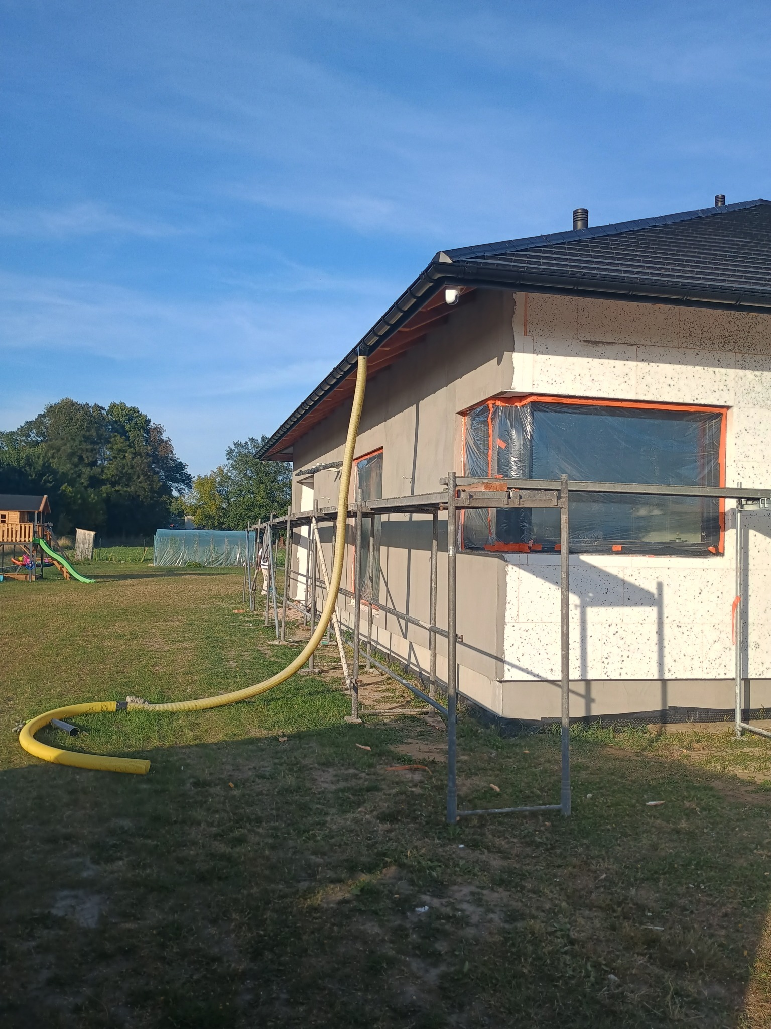 Ocieplanie domu styropianem, widoczne rusztowania i żółty wąż do podawania kleju. Okna zabezpieczone folią. Prace w toku, widok z zewnątrz, słoneczny dzień.