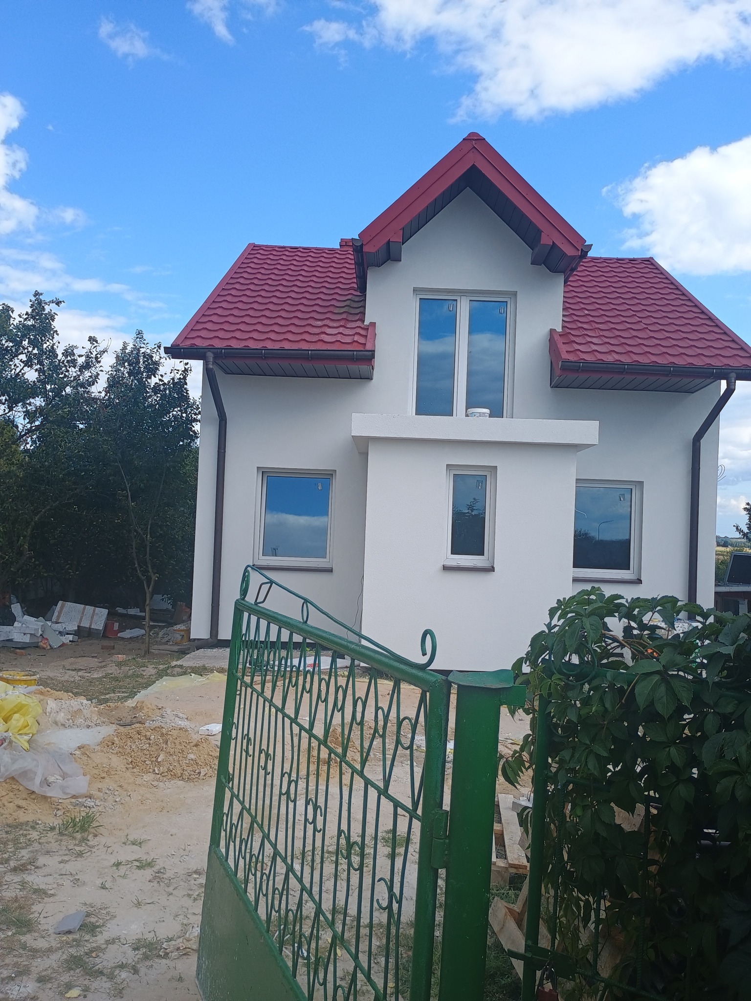 Biały dom z czerwoną dachówką widziany zza zielonego ogrodzenia. Widoczne okna odbijające niebo i materiały budowlane na posesji.