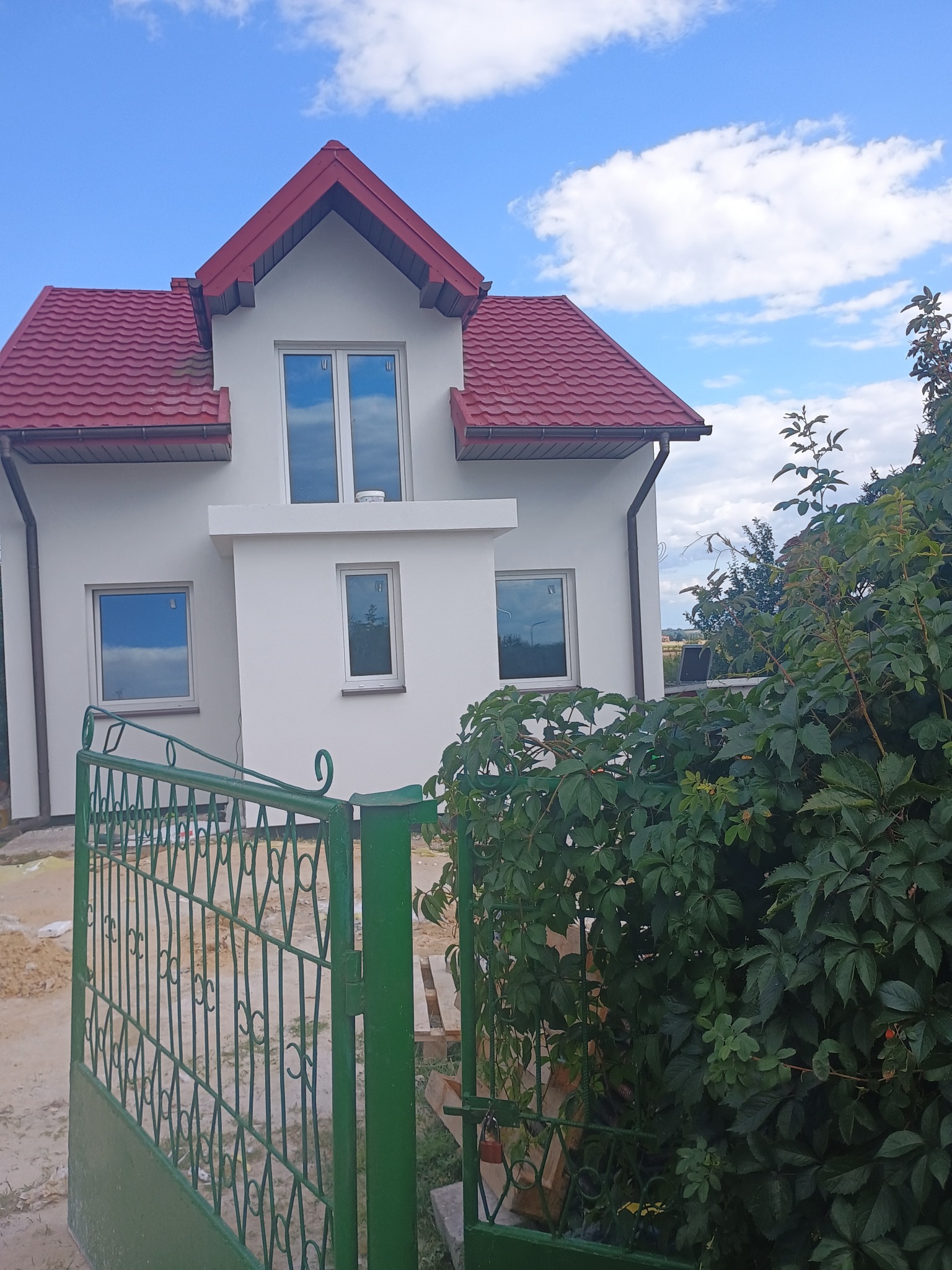 Biały dom z czerwoną dachówką, widziany zza zielonego ogrodzenia i bujnej roślinności. Błękitne niebo z chmurami odbija się w oknach.