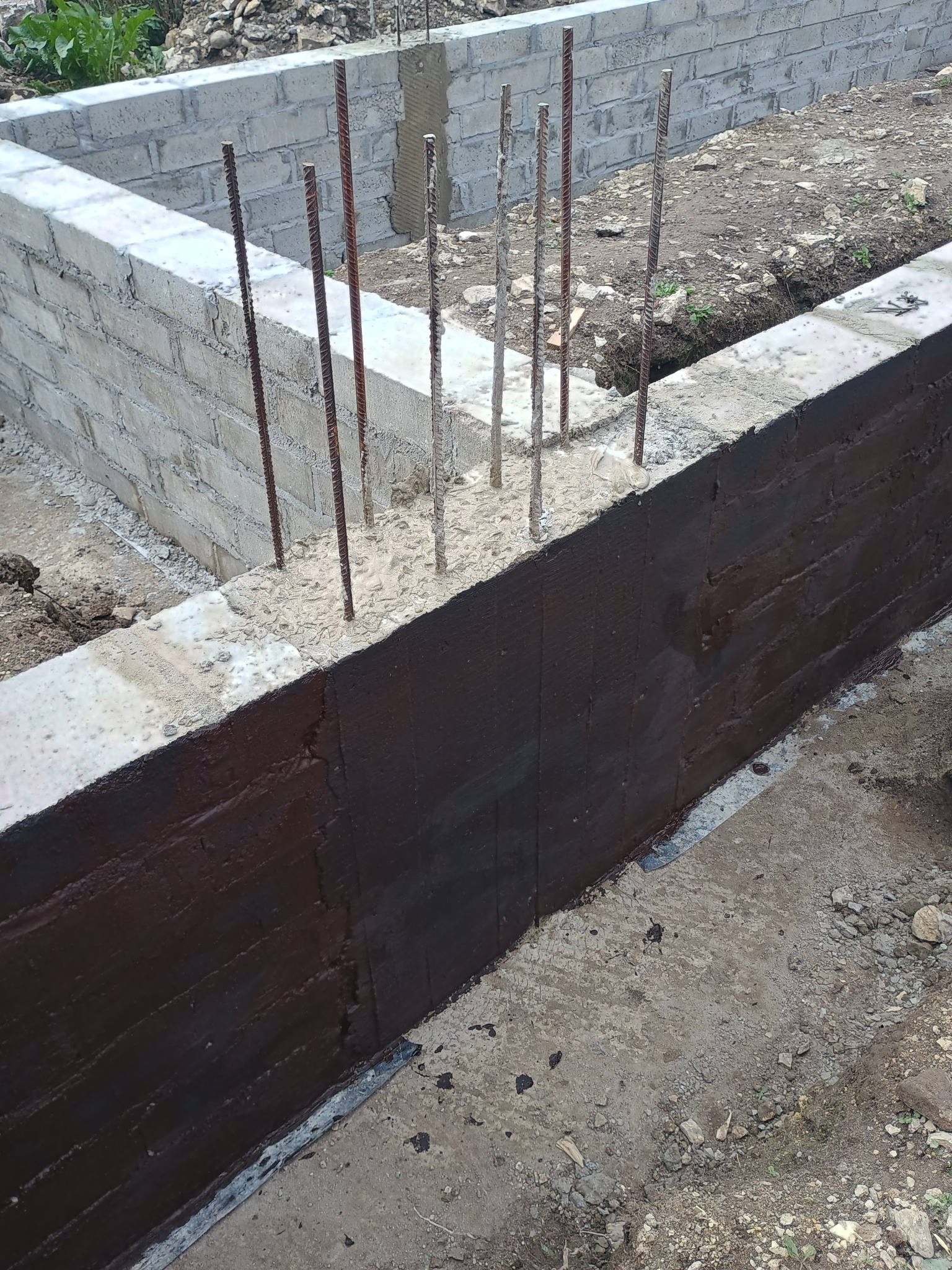 Zabezpieczone fundamenty z betonu komórkowego, pokryte ciemną izolacją przeciwwilgociową. Widoczne wystające pręty zbrojeniowe i fragment terenu budowy.