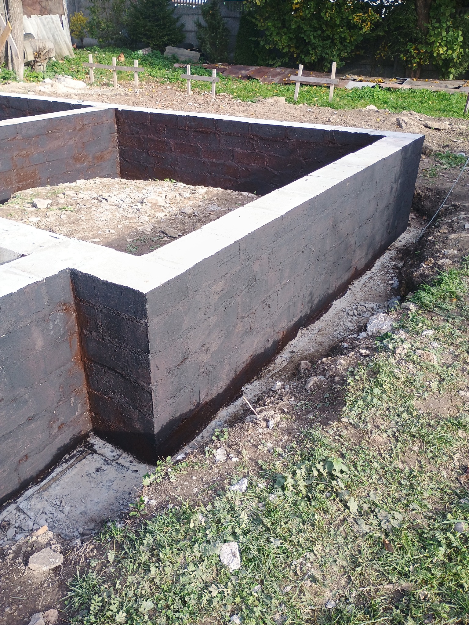 Zabezpieczone, pomalowane fundamenty domu z betonu komórkowego, widoczne z poziomu gruntu. Izolacja przeciwwilgociowa w kolorze czarnym. Otoczenie: trawa, ziemia.