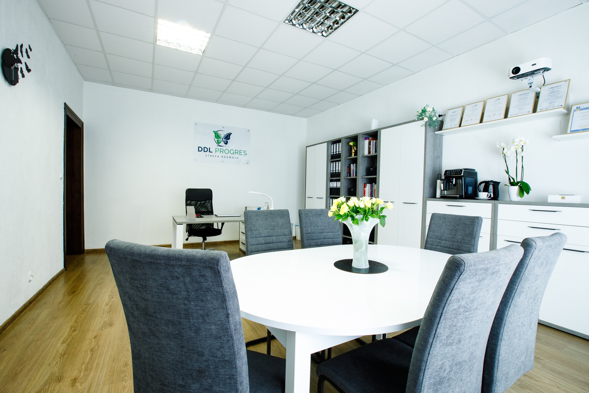 Jasne wnętrze biura z okrągłym stołem, szarymi krzesłami, kwiatami, regałem, certyfikatami i logo firmy DDL PROGRES na ścianie. Nowoczesny i profesjonalny wystrój.