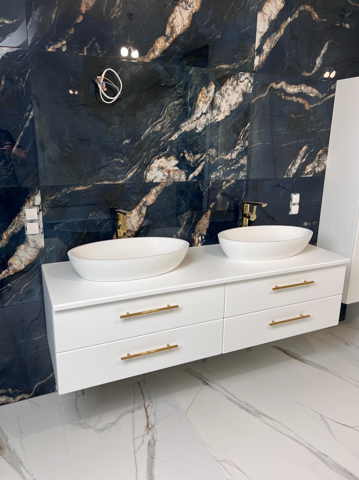 Nowoczesna łazienka z czarną ścianą z marmurowym wzorem, białą szafką z dwiema umywalkami i złotymi bateriami oraz uchwytami, na podłodze białe płytki z szarymi żyłami.
