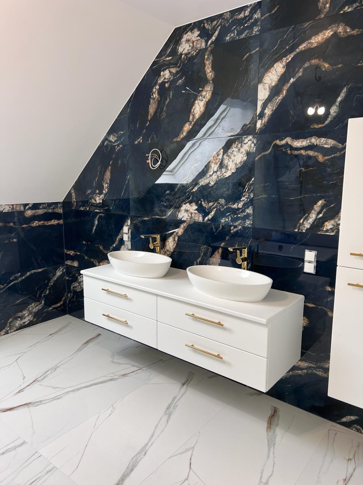 Elegancka łazienka w Oławie z czarnymi płytkami imitującymi marmur na ścianach i białymi płytkami na podłodze. Biała szafka z umywalkami i złotymi detalami.