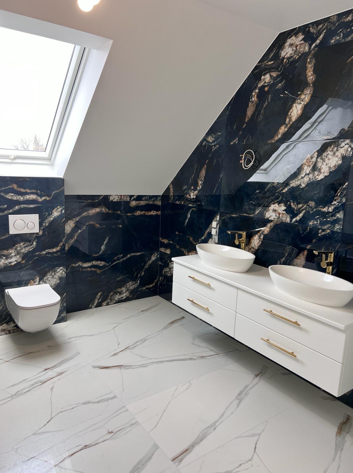 Elegancka łazienka na poddaszu z czarnymi płytkami imitującymi marmur, białą armaturą i podłogą w jasne wzory. Uwagę przykuwają złote detale baterii i uchwytów.