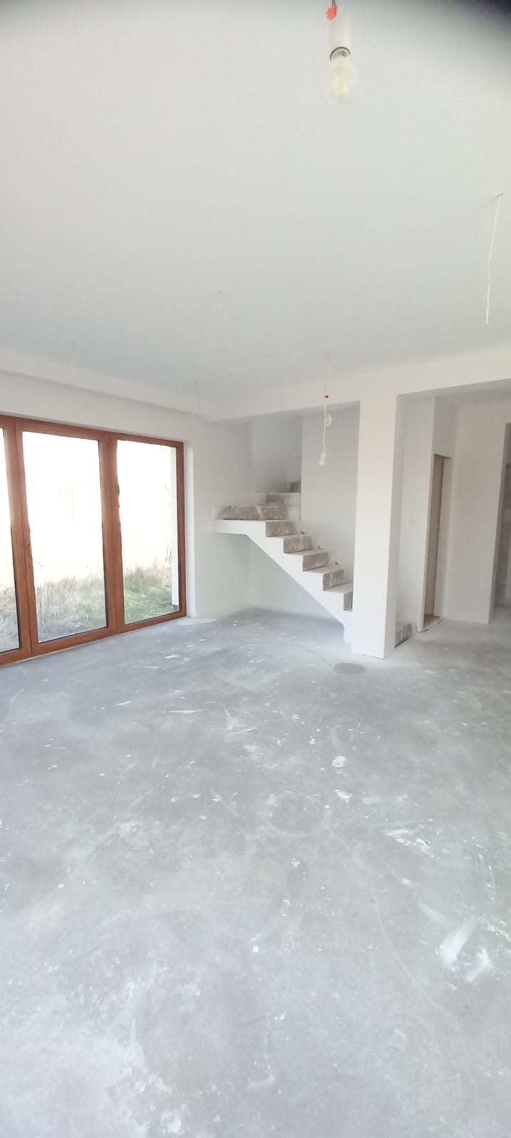 Surowe wnętrze z betonową podłogą, schodami w stanie surowym i dużymi oknami. Widoczne niedokończone wykończenia i wiszące kable.