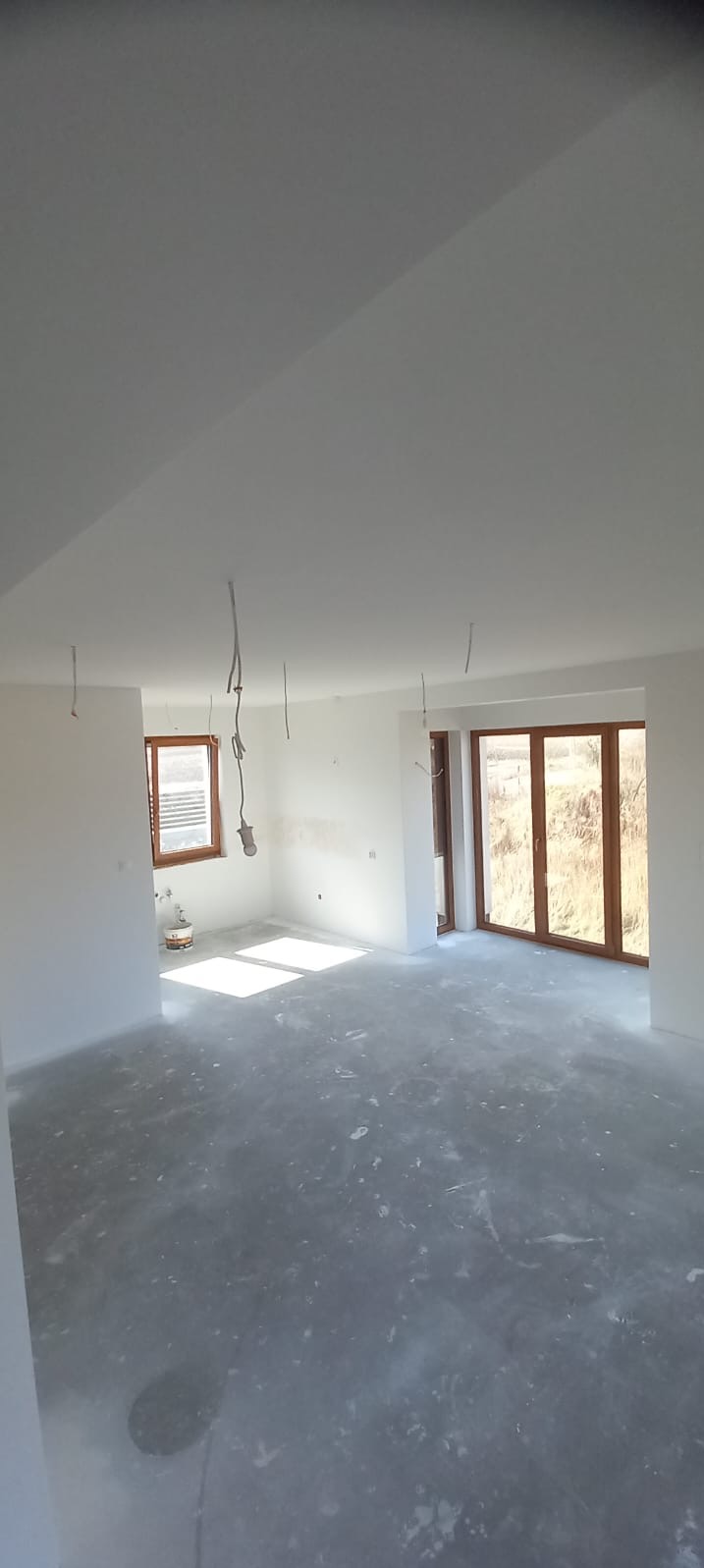 Jasne, przestronne wnętrze po szpachlowaniu, z widocznymi przewodami elektrycznymi i oknami z naturalnym światłem. Świeżo przygotowana podłoga pod dalsze prace wykończeniowe.