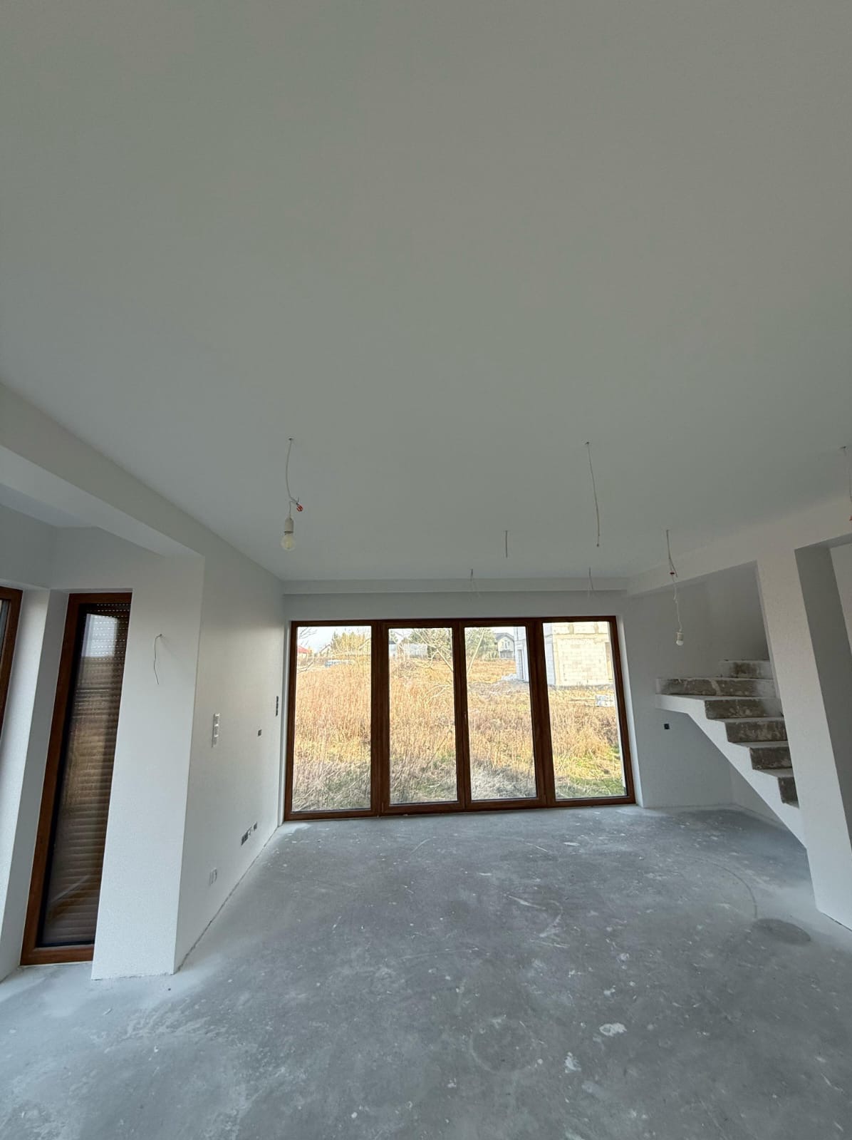 Surowe wnętrze domu w trakcie wykańczania: białe ściany, surowy beton na podłodze, drewniane okna z widokiem na zewnątrz, schody w stanie surowym. Widoczne kable od oświetlenia.