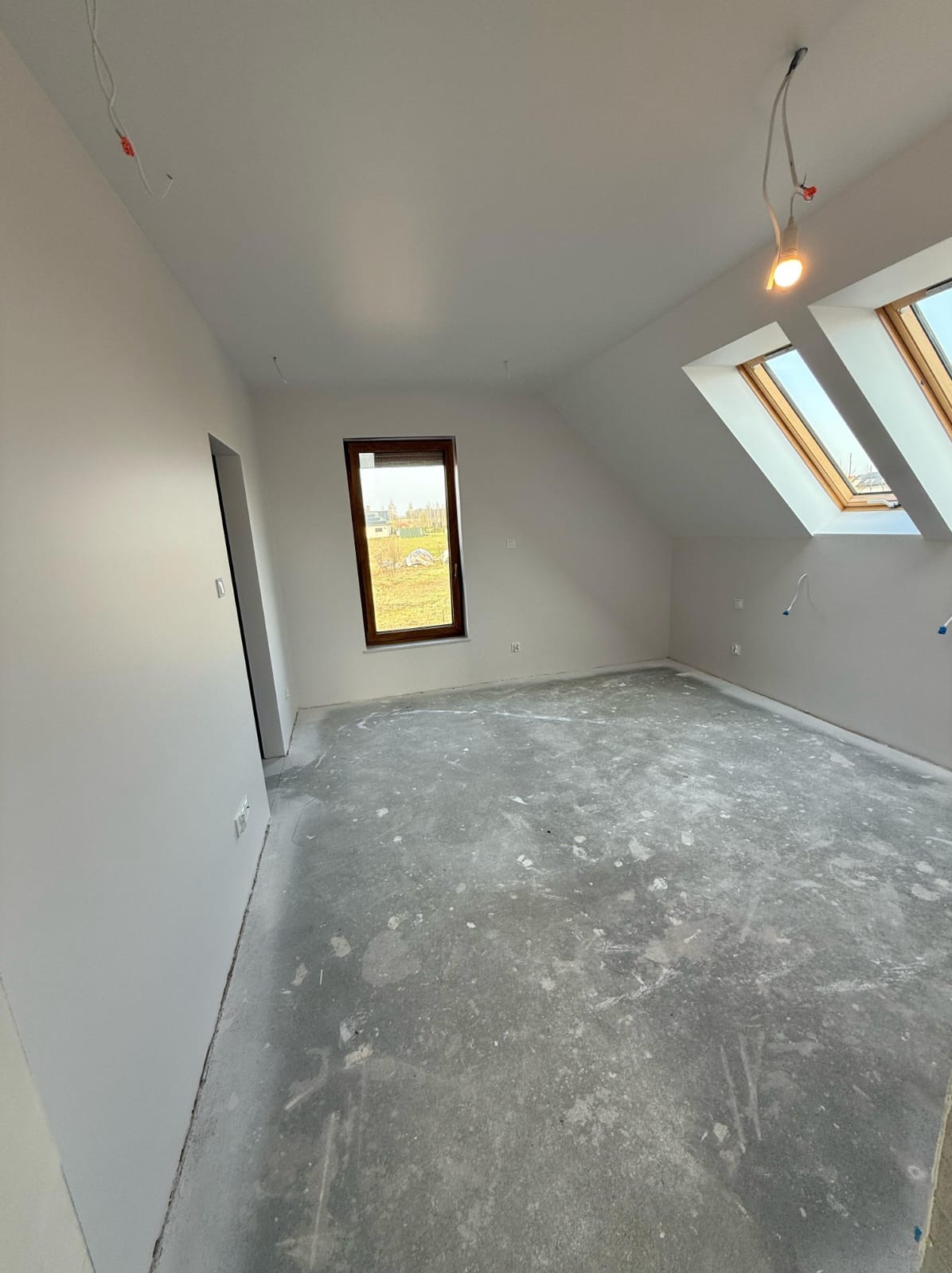 Poddasze w trakcie wykańczania: białe ściany, surowa podłoga, okna dachowe i pionowe. Widoczne kable elektryczne, przygotowane do dalszych prac wykończeniowych.