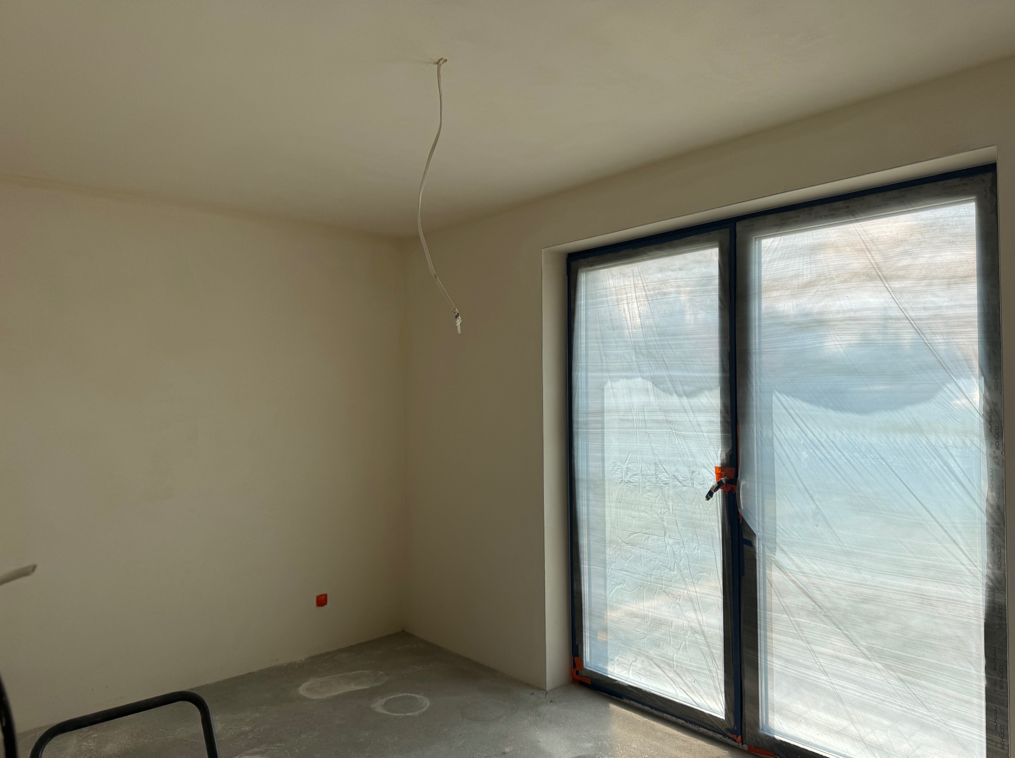 Wnętrze pokoju w trakcie prac wykończeniowych: ściany po szpachlowaniu, zabezpieczone okno folią, widoczny kabel zwisający z sufitu i fragment drabiny malarskiej.