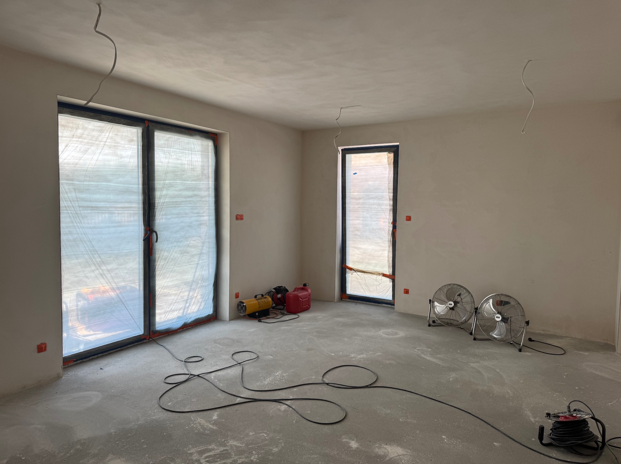 Wnętrze po szpachlowaniu ścian i sufitu, z osłoniętymi oknami, nagrzewnicą i wentylatorami wspomagającymi schnięcie. Widoczne kable na podłodze.