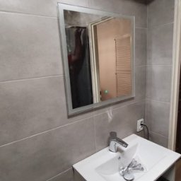 Radosław No - Wykończona łazienka z szarymi płytkami, lustrem z podświetleniem LED i białą szafką pod umywalką. Widoczne narzędzia. Otwór w suficie.