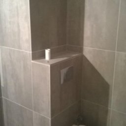 Radosław No - Szara łazienka z podwieszaną toaletą i wnęką wyłożoną płytkami. Minimalistyczny design, nowoczesne wykończenie. Widoczny fragment podłogi i biała rolka papieru.