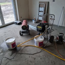 Radosław No - Wnętrze w trakcie malowania maszynowego. Widoczny agregat malarski, wiadro z farbą Atlas, nagrzewnica i drabina. Surowy stan ścian przed ostatecznym wykończeniem.