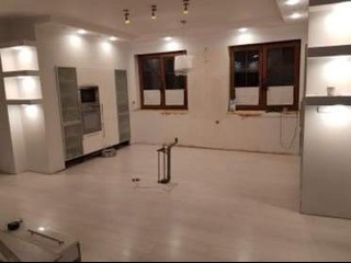 Jasne wnętrze w trakcie remontu: białe ściany, drewniane okna, przygotowana podłoga. Widoczne narzędzia i oświetlenie sufitowe.