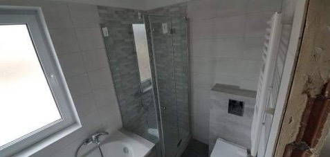 Wykończona łazienka z kabiną prysznicową, umywalką w zabudowie i grzejnikiem. Widoczne szare płytki na ścianach i częściowo odkryta cegła.