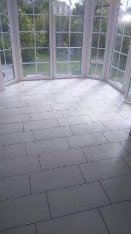 Białe, prostokątne płytki podłogowe w jasnym pomieszczeniu z dużymi oknami wychodzącymi na zieleń. Minimalistyczny design i świeże wykończenie.