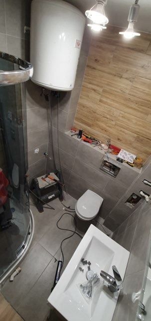 Remont łazienki w trakcie: biały bojler, toaleta, umywalka z baterią, płytki imitujące drewno i szare płytki. Widoczne narzędzia i akcesoria instalacyjne.