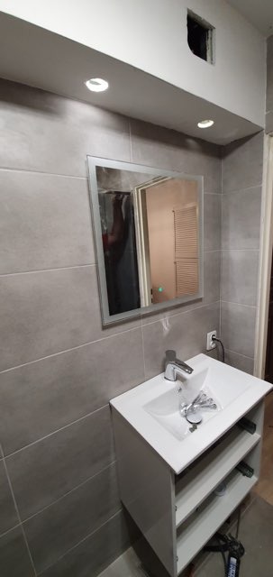 Wykończona łazienka z szarymi płytkami, lustrem z podświetleniem LED i białą szafką pod umywalką. Widoczne narzędzia. Otwór w suficie.