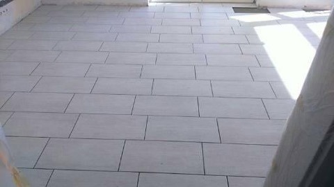 Jasne, prostokątne płytki podłogowe ułożone w regularny wzór, widoczne fugi i fragment okna z promieniami słonecznymi. Minimalistyczny design.