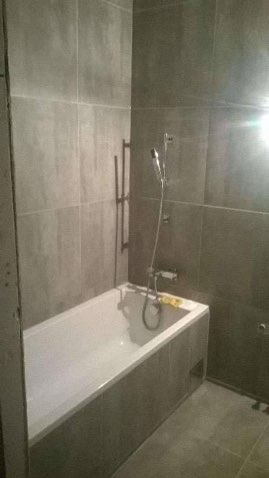 Wykończona łazienka z wanną w zabudowie i szarą glazurą. Minimalistyczny design, widoczny grzejnik drabinkowy i bateria prysznicowa. Nowoczesne wnętrze.