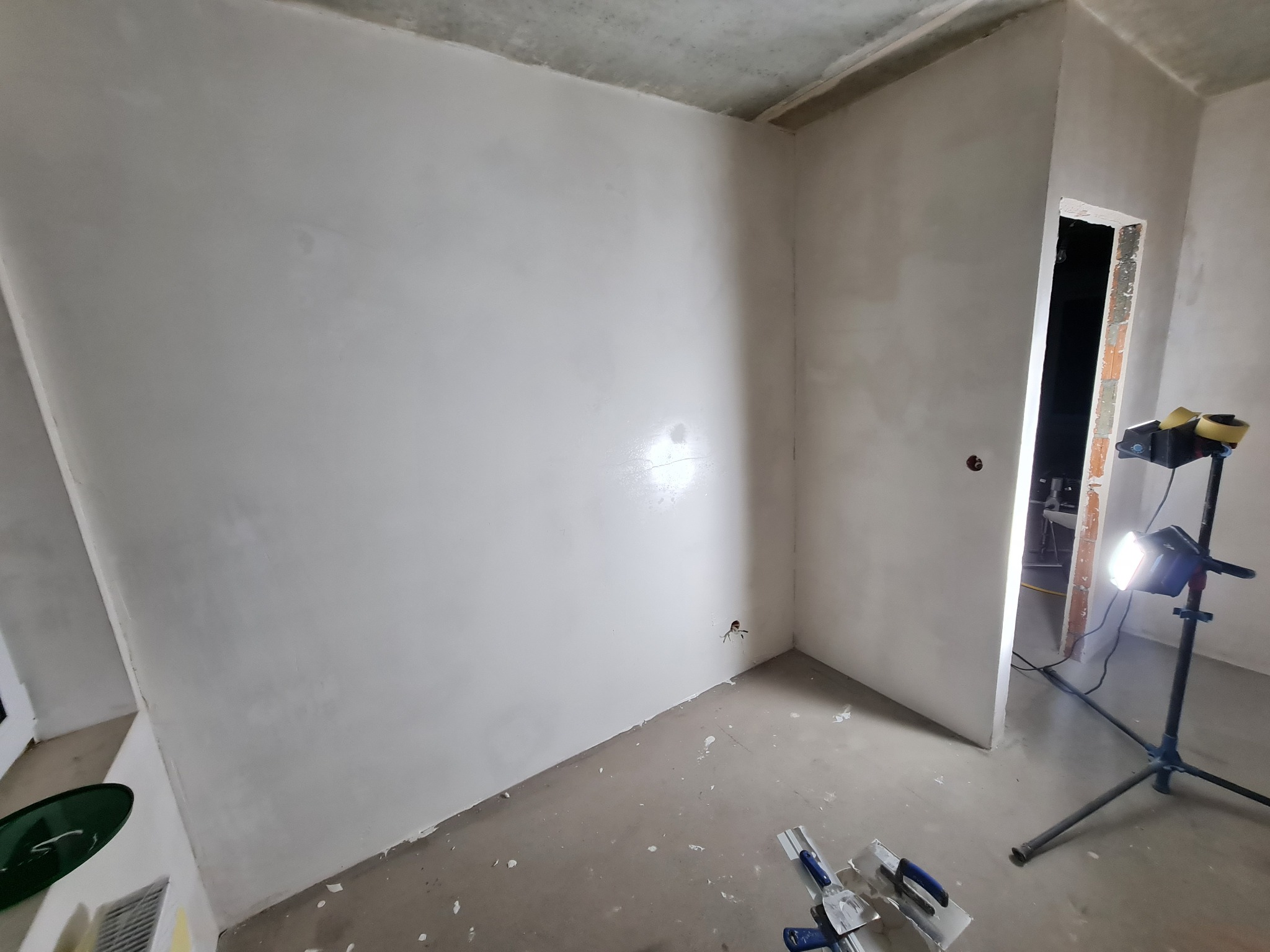 Wnętrze po szpachlowaniu ścian, widoczne narzędzia i lampa oświetlająca. Ściany gładkie, przygotowane do dalszych prac wykończeniowych. W tle otwór drzwiowy.