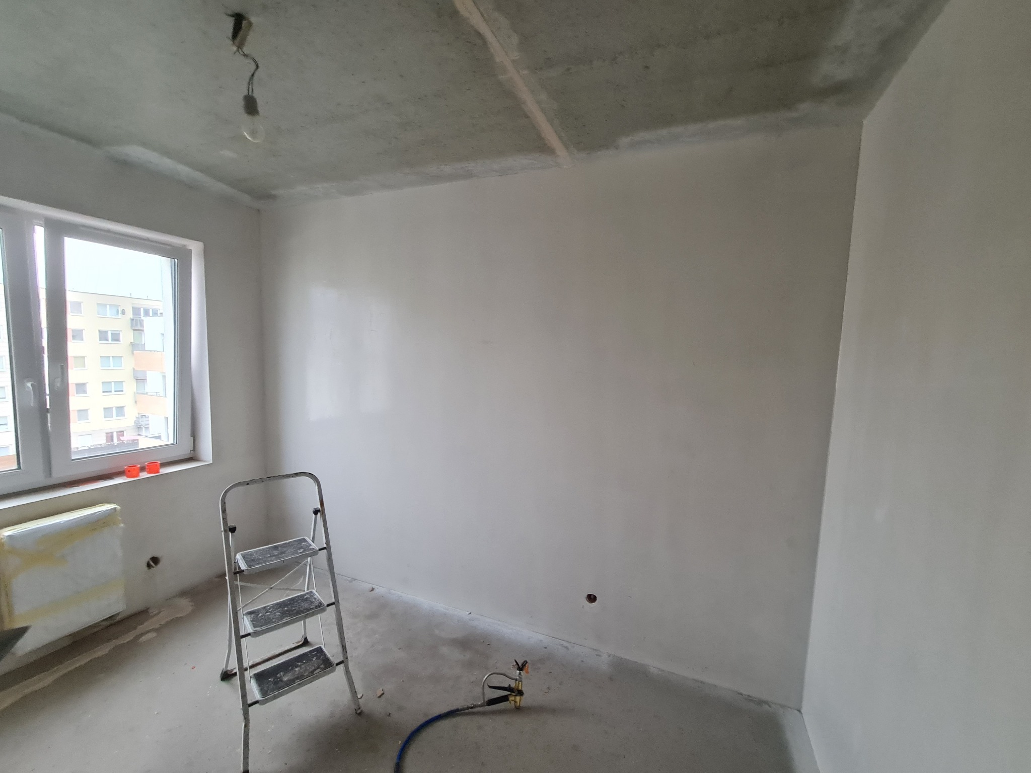 Wnętrze po szpachlowaniu: gładkie ściany, drabina, osłonięty grzejnik, surowy sufit z żarówką. Przygotowanie do malowania, widoczny agregat szpachlarski.