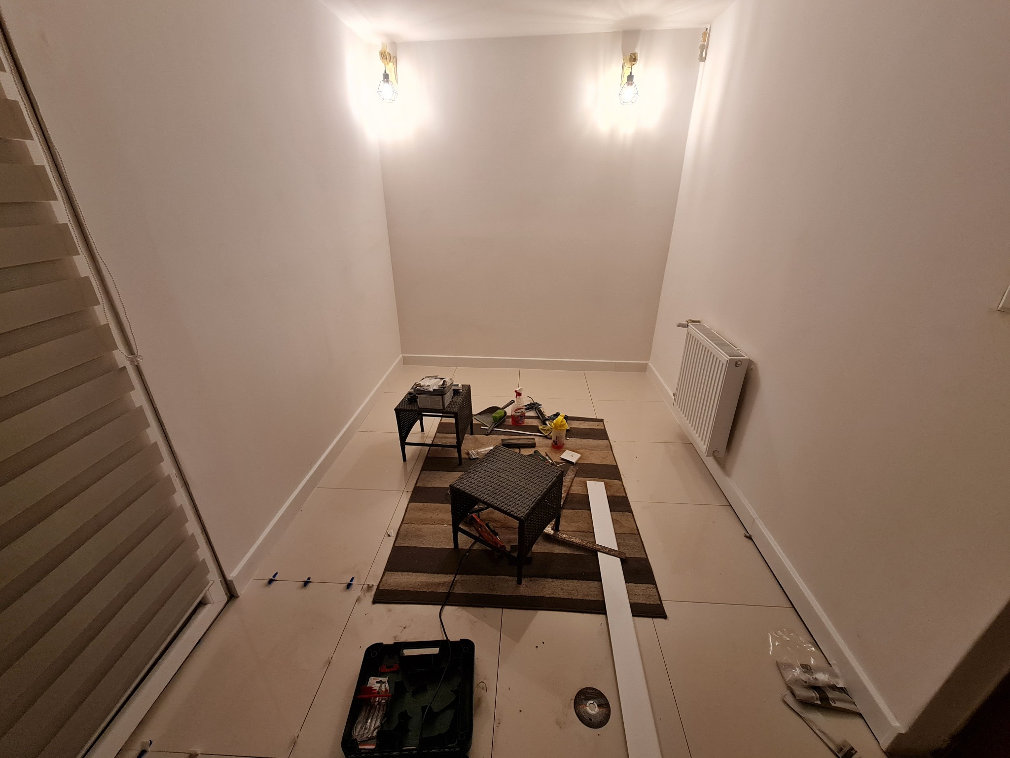 Wnętrze pokoju z narzędziami na dywanie i podłodze, widoczne akcesoria do wykończenia, płytki podłogowe, listwy przypodłogowe i lampy na ścianie.